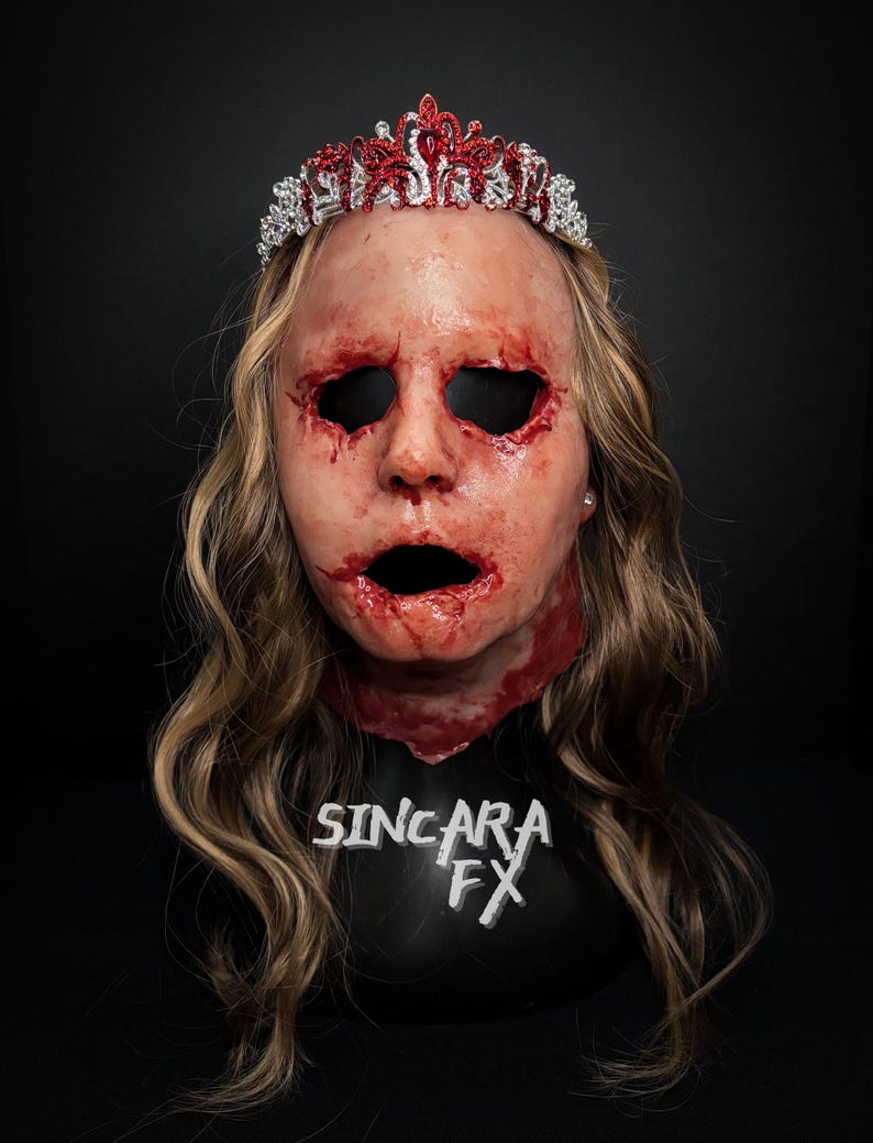 May include: A gruesome Halloween mask with a bloodied face, open mouth, and deep wounds. The mask is adorned with a red and silver tiara and long, wavy blonde hair. The text "SINCARA FX" is visible on the base.