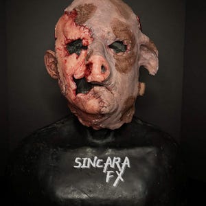 May include: A gruesome mask with a pig-like nose and human characteristics. The mask is a flesh tone with brown patches and visible wounds, stitches, and exposed flesh. The text "SINCARA FX" is printed in white on the black base.