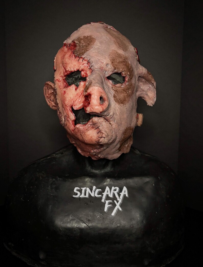 May include: A gruesome mask with a pig-like nose and human characteristics. The mask is a flesh tone with brown patches and visible wounds, stitches, and exposed flesh. The text "SINCARA FX" is printed in white on the black base.