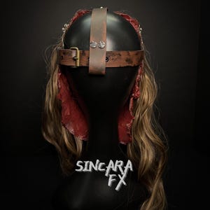 May include: A brown leather headpiece with a buckle and rivets, set against a black background. The headpiece is on a black mannequin head with long, wavy blonde hair and red fabric accents. The text "SINCARA FX" is visible.