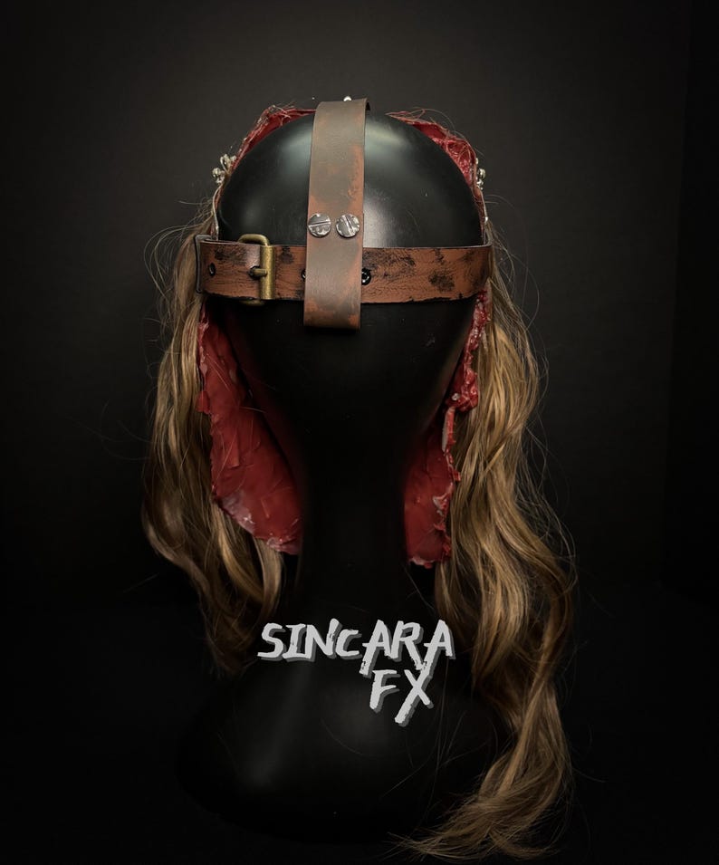 May include: A brown leather headpiece with a buckle and rivets, set against a black background. The headpiece is on a black mannequin head with long, wavy blonde hair and red fabric accents. The text "SINCARA FX" is visible.