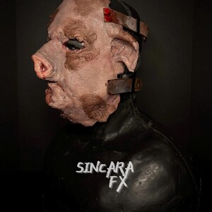 May include: A realistic pig-faced mask with a textured surface and brown markings. The mask has a prominent snout, large ears, and metal straps. The black base features the text "SINCARA FX". The overall design is detailed and unsettling.