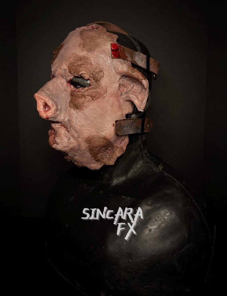 May include: A realistic pig-faced mask with a textured surface and brown markings. The mask has a prominent snout, large ears, and metal straps. The black base features the text "SINCARA FX". The overall design is detailed and unsettling.