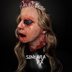 May include: A gruesome mask with a crown, featuring realistic wounds and a gaping mouth. The crown is decorated with red and clear jewels. The mask is set against a black backdrop, with the text "SINCARA FX" visible on the base.