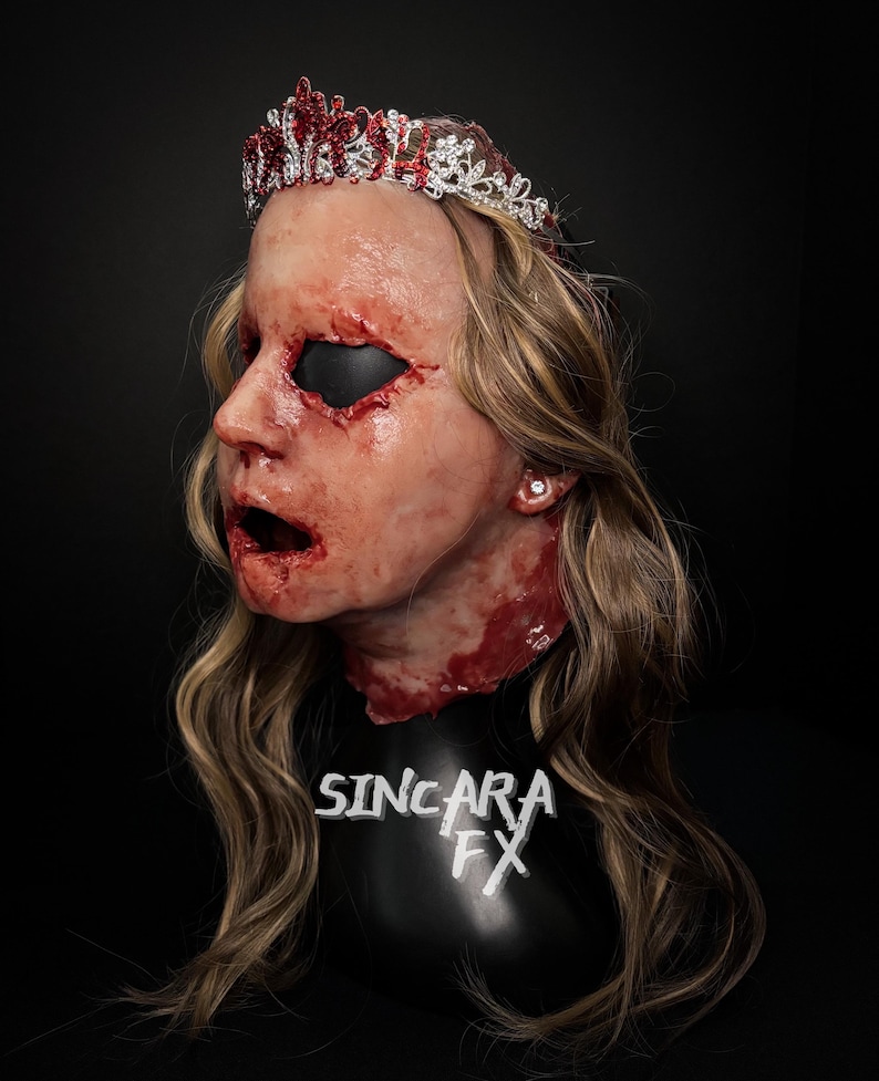 May include: A gruesome mask with a crown, featuring realistic wounds and a gaping mouth. The crown is decorated with red and clear jewels. The mask is set against a black backdrop, with the text "SINCARA FX" visible on the base.