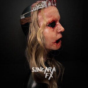 May include: A gruesome mask with blonde hair, a silver and red jeweled crown, and visible wounds. The mask features an open mouth and empty eye sockets. The text "SINCARA FX" is visible on the mask.