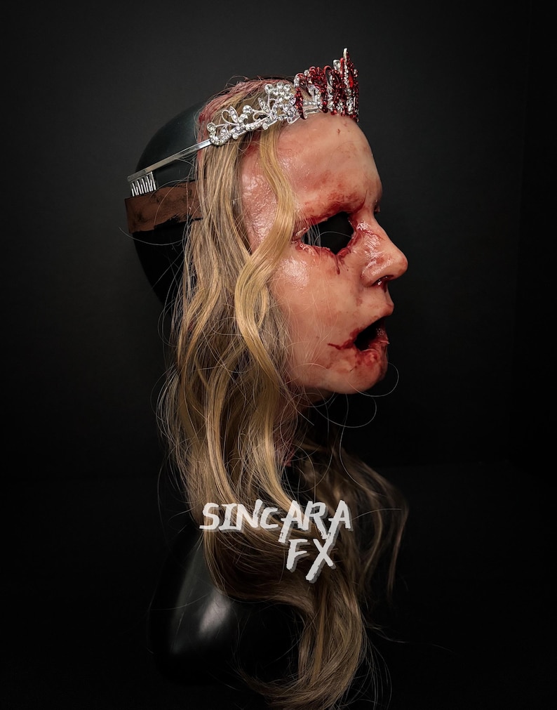 May include: A gruesome mask with blonde hair, a silver and red jeweled crown, and visible wounds. The mask features an open mouth and empty eye sockets. The text "SINCARA FX" is visible on the mask.