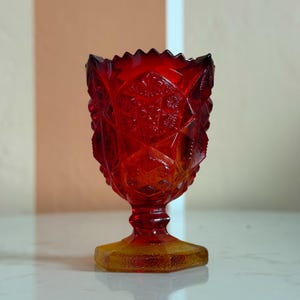 May include: A ruby red, vintage-style glass goblet with a textured, diamond-cut pattern. The goblet has a hexagonal base in amber, a stem, and a scalloped rim. The glass is translucent, allowing light to pass through, and is likely used for serving beverages.