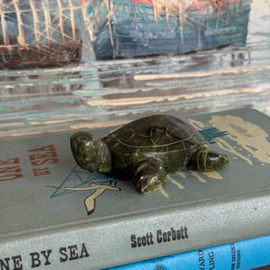 May include: A small, dark green stone turtle figurine rests on a stack of vintage books. The books are titled "One By Sea" and "Captains Courageous." The background features a painting of boats.