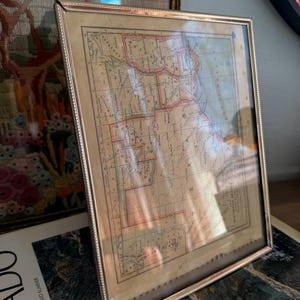 May include: A framed vintage map with a silver-colored metal frame. The map features a detailed layout with red and brown accents. The frame has a beaded edge. The map is displayed on top of a book with the word "COLORADO" visible.