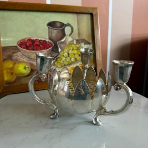May include: Silver-colored metal candle holder with three candle cups and a decorative floral design. The candle holder has curved handles and sits on three small feet. A framed still life painting is in the background.