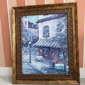 May include: A framed painting depicts a European cafe scene. The artwork features a building with a striped awning, outdoor seating, and a street lamp. The frame is ornate with a gold finish.