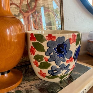 May include: A cream-colored ceramic planter featuring a hand-painted floral pattern with large blue flowers, small red blossoms, and green leaves. The cylindrical vessel has a smooth finish and a wide opening, showcasing a vibrant botanical design.