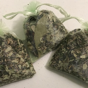 May include: Three small, mesh bags filled with dried herbs and leaves. The bags are tied with light green ribbons. The contents of the bags appear to be a mix of green and purple dried herbs and leaves.
