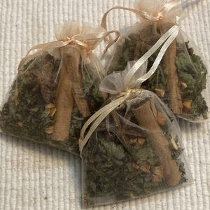 May include: Three small, sheer mesh bags filled with dried herbs and spices. Each bag contains green leaves, small orange pieces, and a cinnamon stick. The bags are tied with a light brown ribbon.