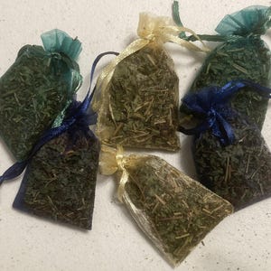 May include: Five small rectangular organza drawstring pouches filled with dried green herbs. The pouches feature sheer fabric in shades of teal, gold, and dark blue, each secured with matching ribbon ties at the top to contain the loose plant matter.