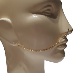 May include: A gold-colored nose ring with a chain that connects to an earring. The jewelry is displayed on a mannequin head. The chain is delicate and hangs from the nose to the ear.