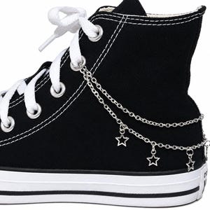 Puede incluir: Una zapatilla de lona negra de caña alta con cordones blancos y ojales de metal plateado. Un accesorio de doble cadena plateada está sujeto a los ojales, adornado con tres pequeños dijes en forma de estrella calada en el lateral.