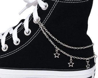 May include: A black high-top canvas sneaker featuring white laces and silver-toned metal eyelets. A double-layered silver chain accessory is attached to the eyelets, adorned with three small, open-frame star charms hanging along the side.