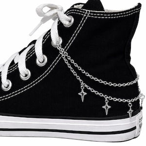 May include: Black high-top canvas sneaker with white laces and a white sole. The sneaker is adorned with a silver chain and star charms, adding a decorative element to the shoe.