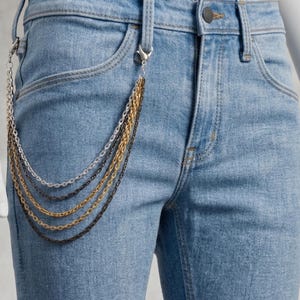 May include: Light blue denim jeans featuring a multi-layered metal chain accessory attached to the belt loops. The accessory consists of five distinct draped chains in varying metallic finishes, including silver, gold, and dark gunmetal tones.