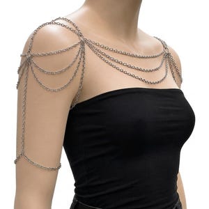 May include: A silver-toned metal body chain accessory featuring multiple draped link chains that extend across the shoulders, chest, and upper arm. The design includes circular connectors that join the layered chains into a cohesive structure.