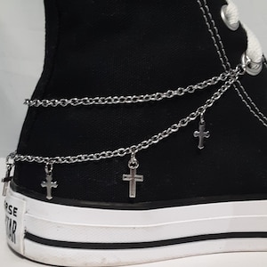 Puede incluir: Zapatillas negras de caña alta con una cadena plateada y dijes de cruz atados a los cordones.