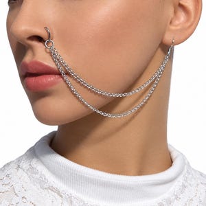 May include: A silver-toned nose-to-ear chain jewellery piece consisting of a circular nose ring connected to two delicate, draped metal link chains that extend to a single earring hook, creating a layered look across the cheek area.