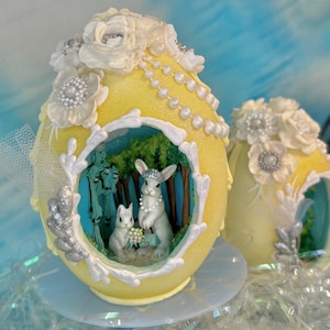 May include: A yellow decorative egg with a scene of two rabbits in a forest. The egg is adorned with white floral accents, pearls, and silver embellishments. The egg is on a light blue surface.