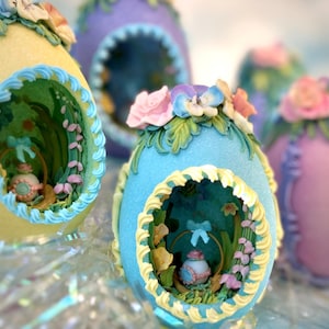 May include: A collection of pastel-colored Easter egg ornaments. Each egg features a decorative opening revealing a miniature scene with a basket and flowers. The eggs are adorned with floral embellishments in shades of pink, blue, and yellow.