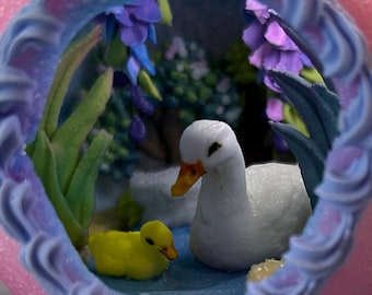 May include: A miniature diorama featuring a white duck and a yellow duckling in a pond setting. The scene is framed by a decorative border with blue and purple accents, and includes floral elements.