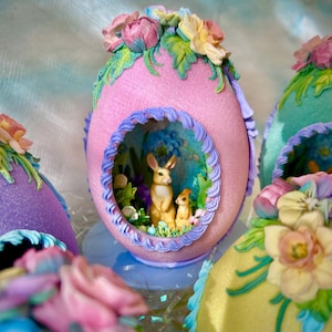 May include: A pink Easter egg with a decorative opening revealing two small rabbit figurines in a garden setting. The egg is adorned with pastel-colored flowers and foliage. Other decorated eggs are visible in the background.