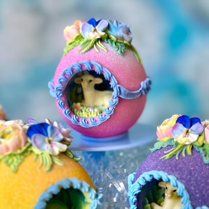 May include: Three decorated Easter eggs with pastel colors and intricate details. Each egg features a small lamb figurine inside a cutout, surrounded by floral accents. The eggs are displayed on a tiered stand.