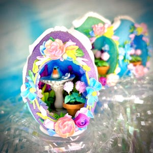 May include: A decorative egg-shaped diorama with a purple exterior, adorned with colorful flowers and a miniature bird bath. Inside, a blue bird perches near a small potted plant. The background features two additional egg dioramas with similar floral arrangements.