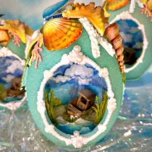 May include: A teal egg-shaped ornament featuring a cutout window revealing a miniature scene with a brown boat, green grass, and a blue sky. The exterior is decorated with various seashells, a starfish, and white textured coral-like accents.