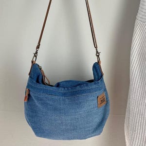 Op de afbeelding: Een blauwe denim schoudertas met een bruine leren band en een klein bruin leren patch met tekst.