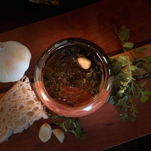 May include: Overhead shot of a glass bowl filled with olive oil, herbs, and a clove of garlic. A whole garlic bulb, bread, and sprigs of fresh herbs are arranged around the bowl on a wooden cutting board. A rustic food preparation.