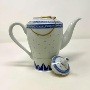 May include: White porcelain teapot with blue geometric and leaf patterns. The teapot has a curved handle and spout, with a lid resting beside it. A gold-colored chain connects the lid to the body. The teapot is a traditional style.