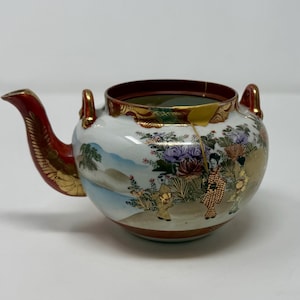 May include: A Japanese-style ceramic teapot with a red handle and spout, and gold accents. The white body features a painted scene with figures, flowers, and landscapes. The rim is dark red with gold trim.