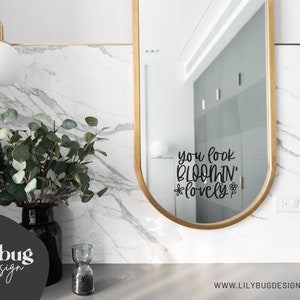 K&ouml;nnte beinhalten: Ein ovaler Spiegel mit goldenem Rahmen und einem schwarzen Vinyl-Aufkleber mit der Aufschrift "you look bloomin' lovely" mit einer kleinen Blumengrafik auf jeder Seite des Wortes "lovely".