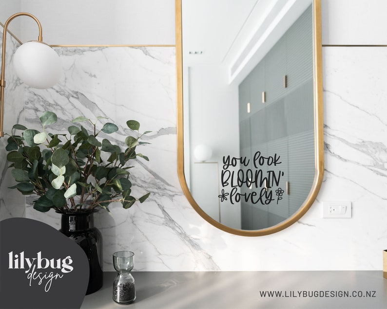 K&ouml;nnte beinhalten: Ein ovaler Spiegel mit goldenem Rahmen und einem schwarzen Vinyl-Aufkleber mit der Aufschrift "you look bloomin' lovely" mit einer kleinen Blumengrafik auf jeder Seite des Wortes "lovely".
