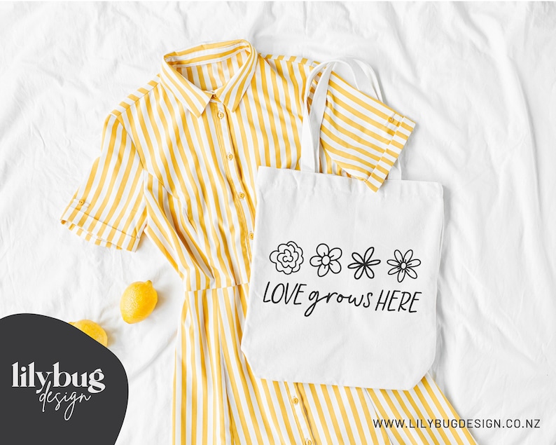 Op de afbeelding: Witte tote bag met zwarte tekst die "LOVE grows HERE" zegt en vier eenvoudige bloemillustraties. De tas ligt op een wit oppervlak met een geel-wit gestreepte jurk en twee citroenen.