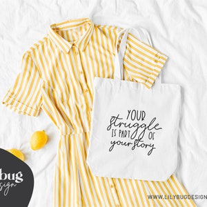 May include: White tote bag with black text that reads "Your struggle is part of your story". The bag is lying on a white surface with a yellow striped shirt and lemons.