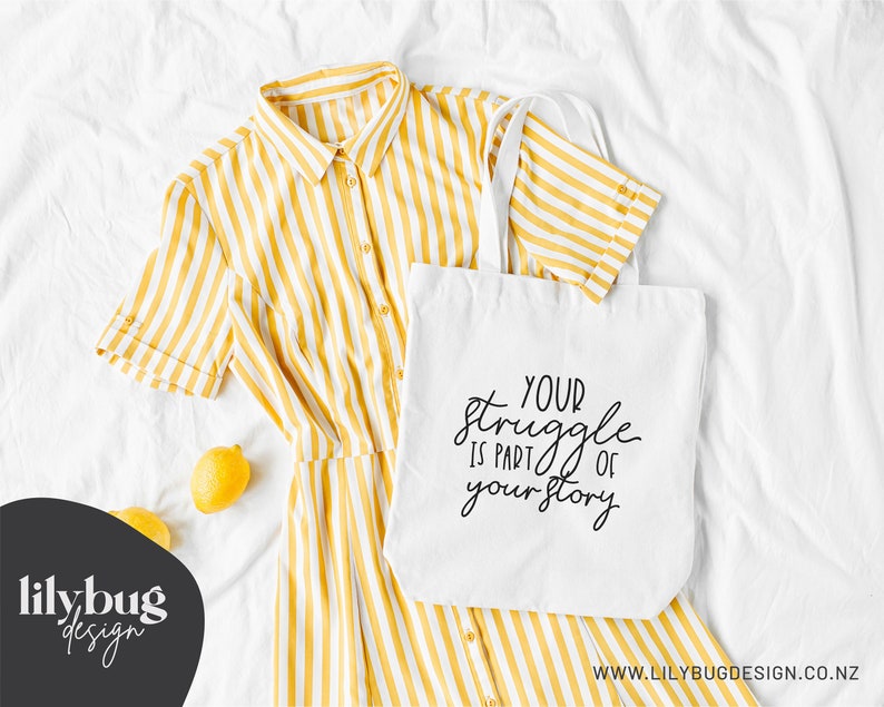 May include: White tote bag with black text that reads "Your struggle is part of your story". The bag is lying on a white surface with a yellow striped shirt and lemons.