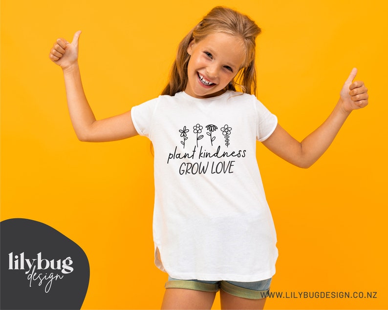 Op de afbeelding: Een wit T-shirt met een zwarte afbeelding waarop "plant kindness grow love" staat, met een rij kleine bloemen boven de tekst.