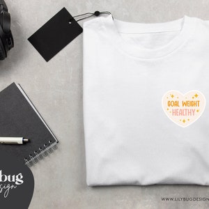 K&ouml;nnte beinhalten: Wei&szlig;es T-Shirt mit einem rosafarbenen herzf&ouml;rmigen Grafik, auf dem "Goal Weight - Healthy" in goldenen Buchstaben mit goldenen Glitzerakzenten steht.