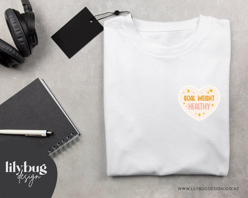 K&ouml;nnte beinhalten: Wei&szlig;es T-Shirt mit einem rosafarbenen herzf&ouml;rmigen Grafik, auf dem "Goal Weight - Healthy" in goldenen Buchstaben mit goldenen Glitzerakzenten steht.