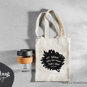 May include: A white tote bag with black handles and the text "My brain has too many tabs open" printed on the front. The bag is sitting on a gray surface next to a reusable coffee cup.