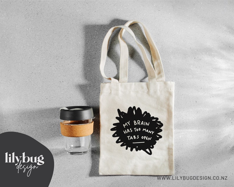 May include: A white tote bag with black handles and the text "My brain has too many tabs open" printed on the front. The bag is sitting on a gray surface next to a reusable coffee cup.