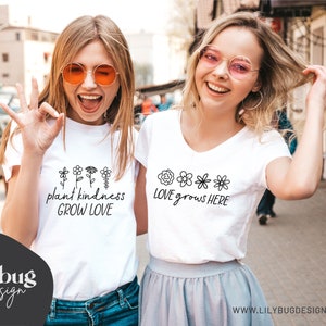 Op de afbeelding: Twee vrouwen dragen witte T-shirts met verschillende bloemenontwerpen en tekst. Het eerste shirt zegt "plant kindness GROW LOVE" en het tweede shirt zegt "LOVE grows HERE".